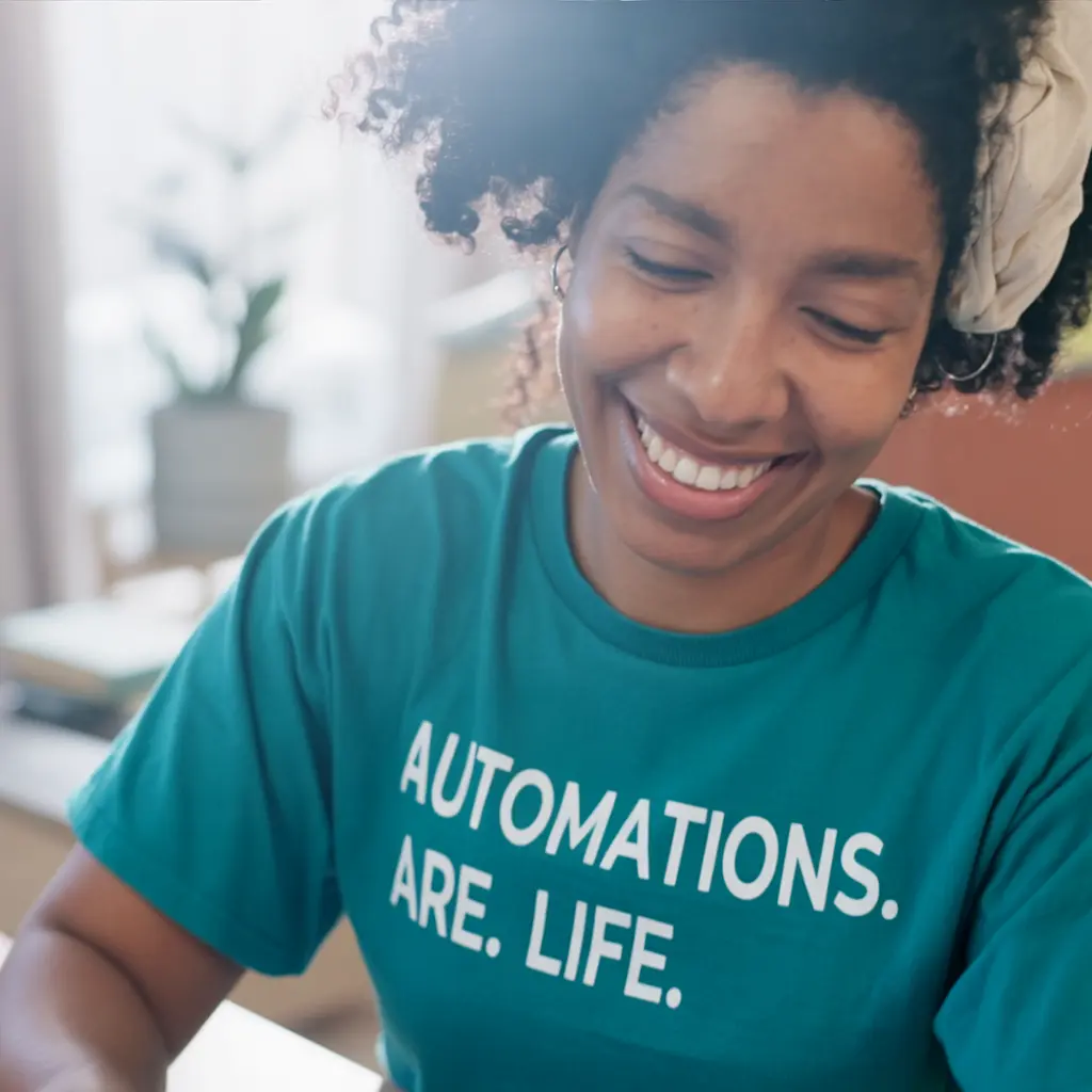smiling-black-woman-wearing-teal-automations-are-life-shirt-working-at-desk-in-bright-room-coaching-that-clicks-ai-automation-lifestyle