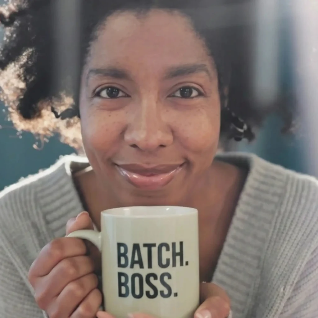 Woman smiling softly while holding a light-colored mug that says “BATCH. BOSS.” in bold black letters. She’s wearing a cozy gray sweater with natural light highlighting her face, capturing the calm confidence of coaching that clicks.