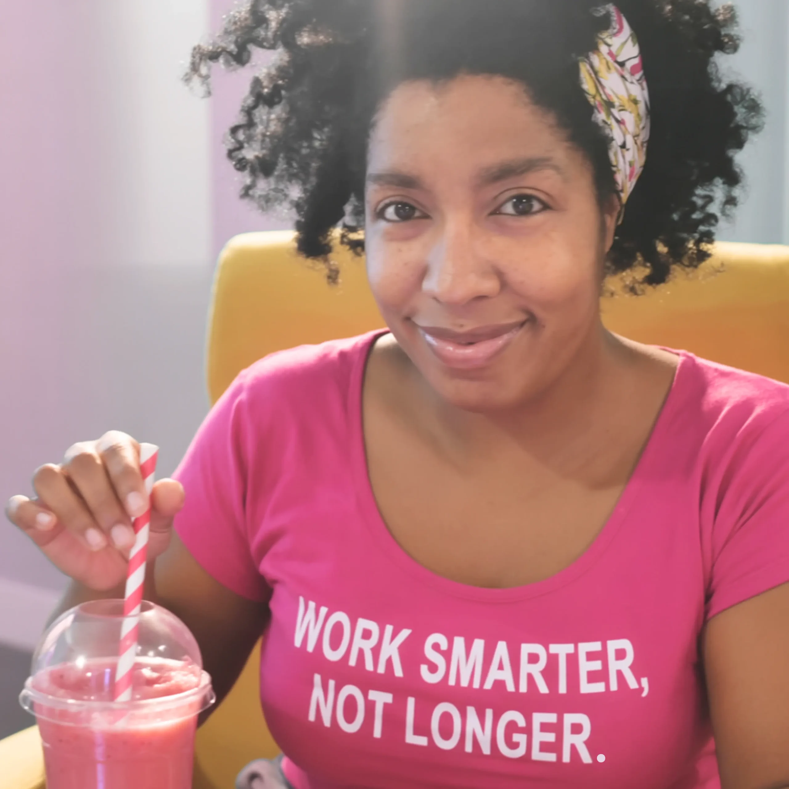 Smiling woman sitting in a yellow chair, holding a pink smoothie with a striped straw, wearing a bright pink shirt that says “WORK SMARTER, NOT LONGER.” Her relaxed confidence and vibrant style reflect the energy of coaching that clicks.